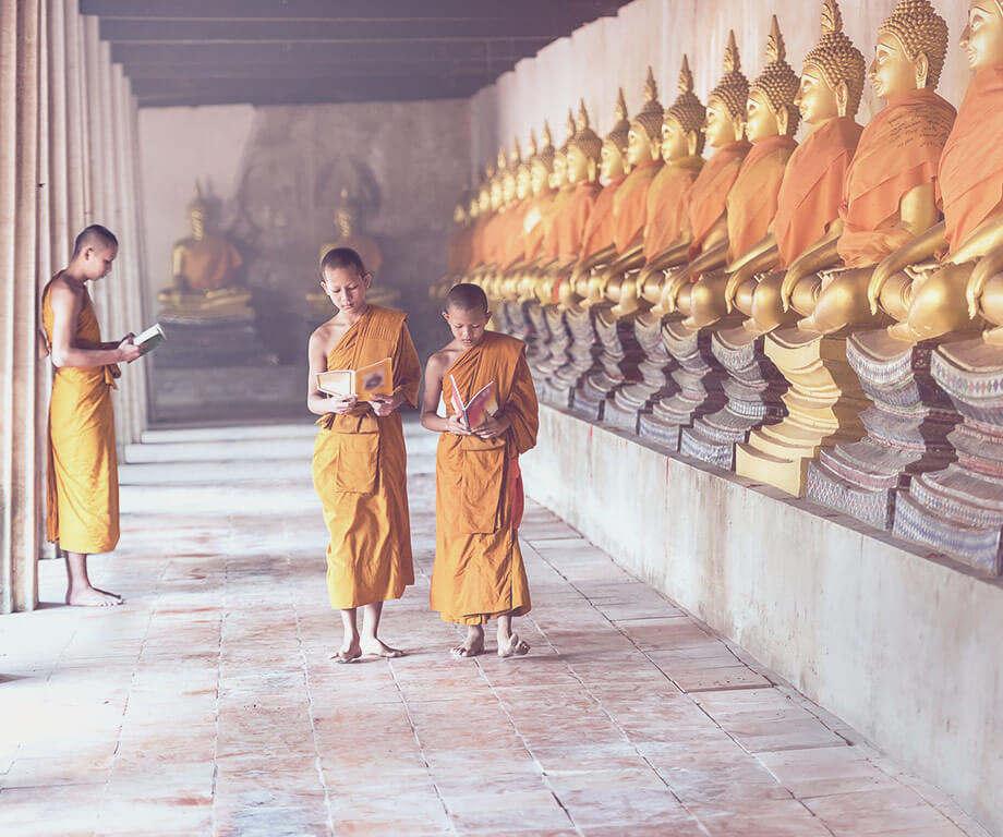 Two young Buddhist students next to a row of statues reading a book. In the background a slightly older student, also reading a book