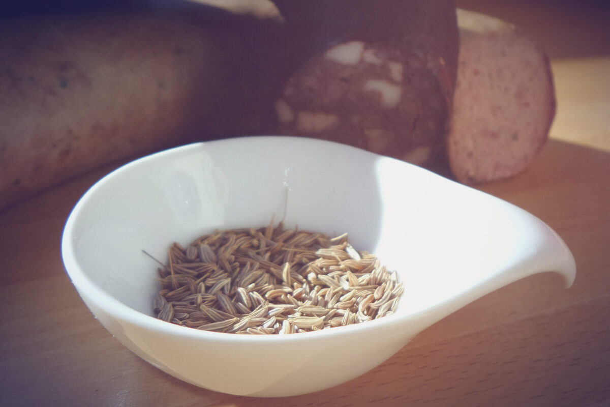 Caraway seeds in a small porcelain bowl, sliced sausage in the background (blurred)
