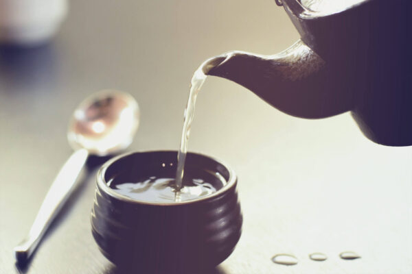Small round tea cup, tea is poured from a clay pot. In the background a large spoon.
