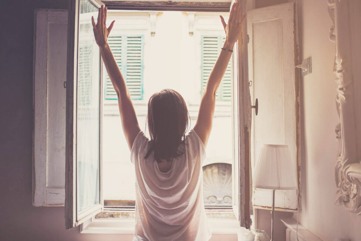 Woman stretching her hands up, photographed from behind in front of open window