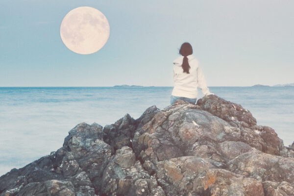 Woman photographed from behind sitting on rocks on beach watching full moon by day